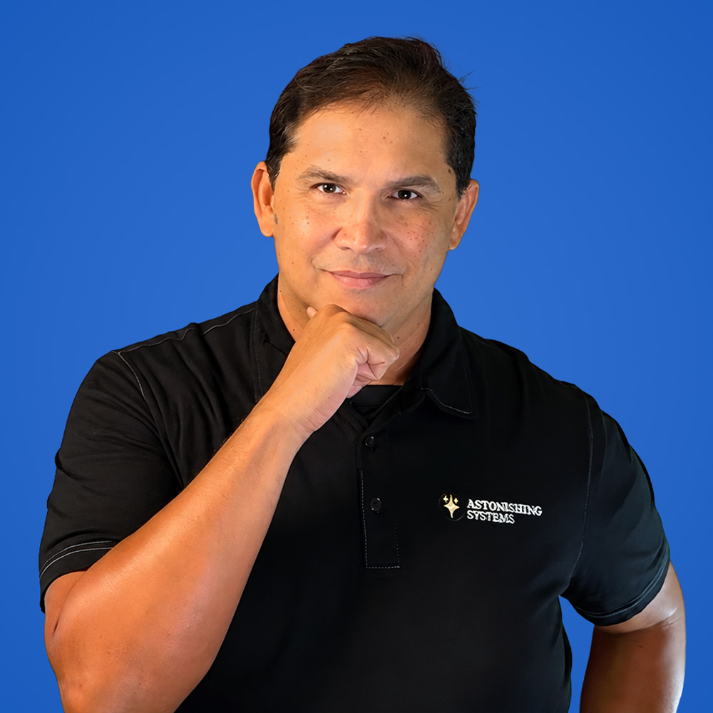Portrait of a man in a black polo shirt with the Astonishing Systems logo, resting his chin on his hand against a solid blue background.