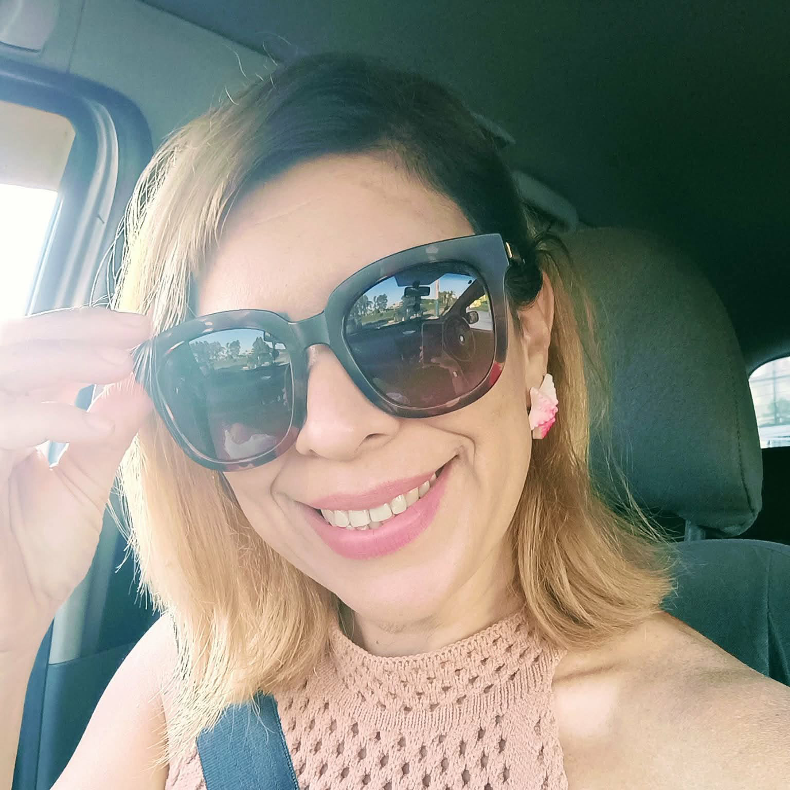 Smiling woman wearing oversized sunglasses in a car, with a seatbelt across her chest and pink knit top.