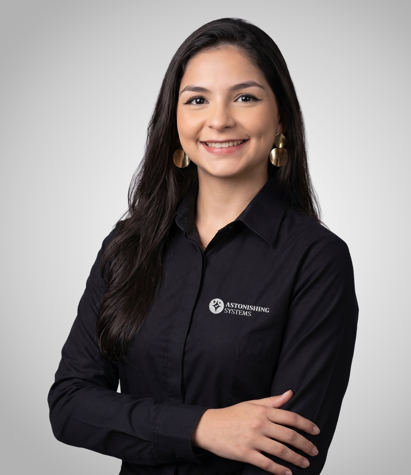 Portrait of a smiling woman with long dark hair, wearing a black collared shirt featuring the Astonishing Systems logo, arms crossed against a gray gradient background.