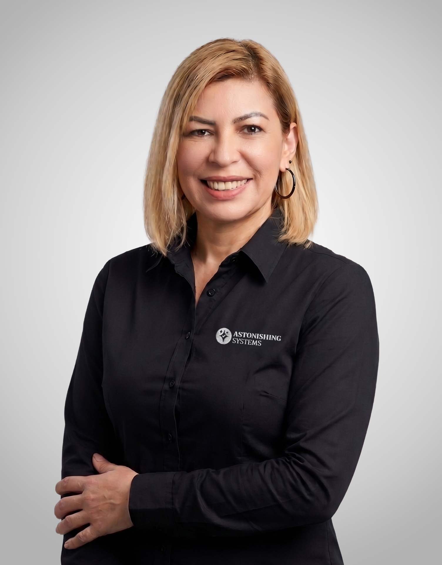 Portrait of a smiling woman in a black shirt with the Astonishing Systems logo on the chest, arms crossed, against a gray gradient backdrop.