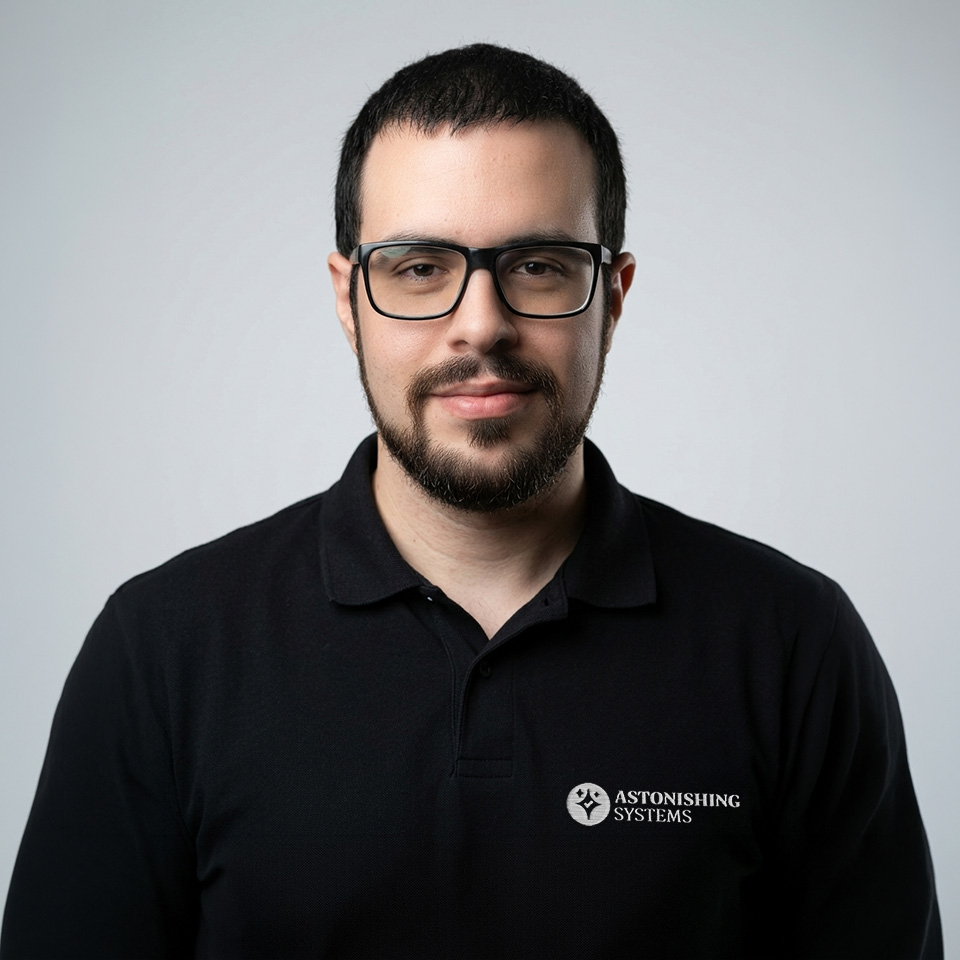 Portrait of a man with short dark hair and beard wearing glasses and a black polo shirt with the Astonishing Systems logo.