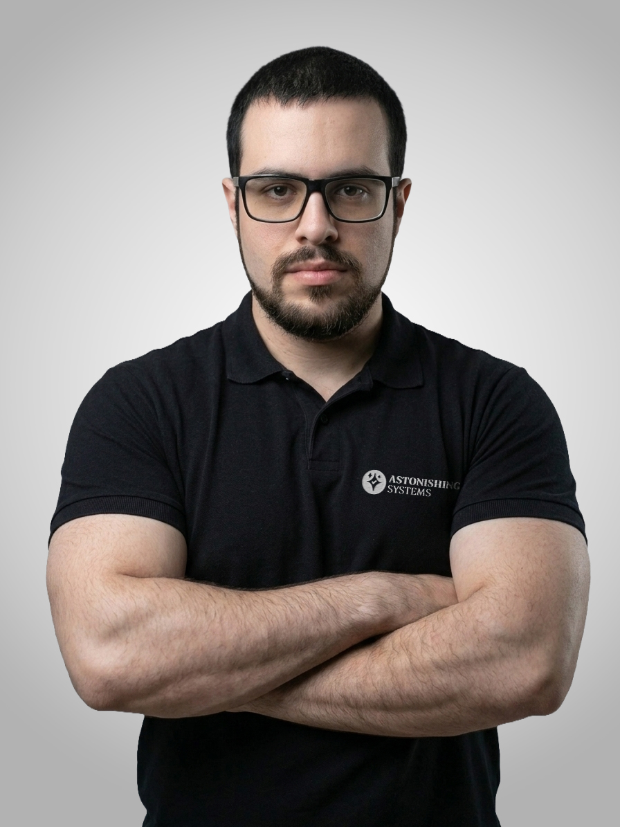 Portrait of a man with short dark hair and glasses, arms crossed, wearing a black polo with an Astonishing Systems logo.